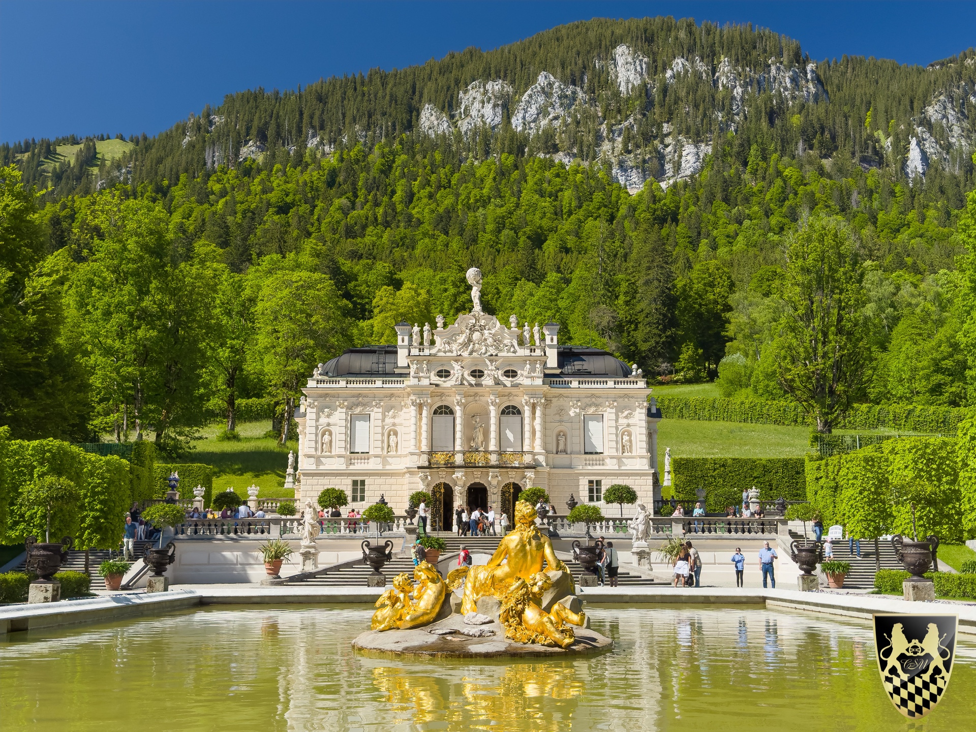 Elegant palace with a fountain in front, set against a mountainous, forested backdrop.