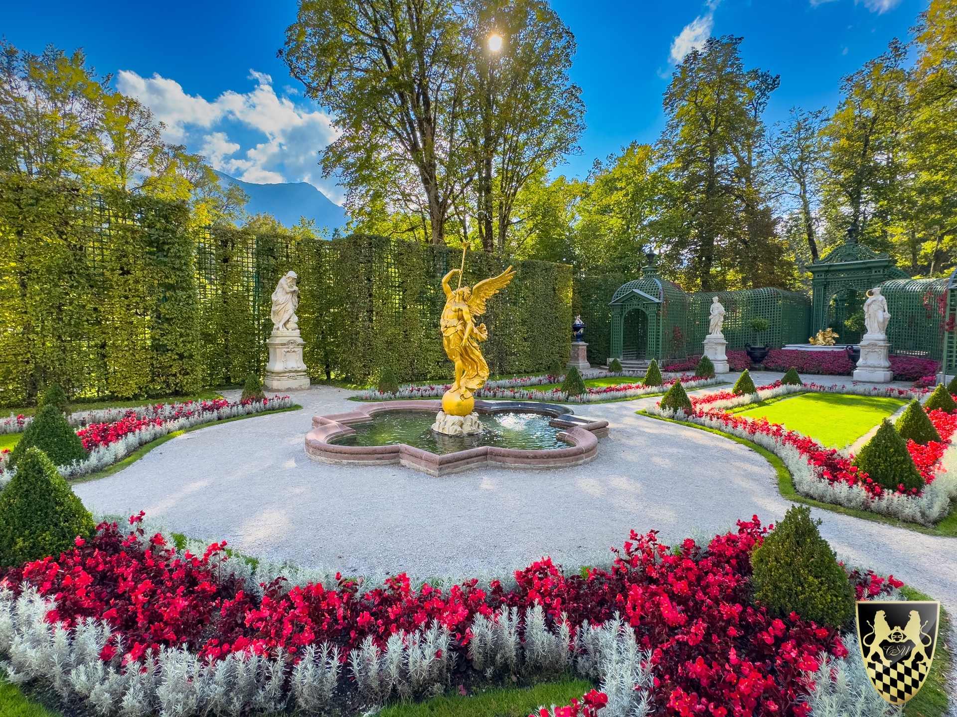 Ornate garden with central golden statue, colorful flowers, and hedges under a bright blue sky.