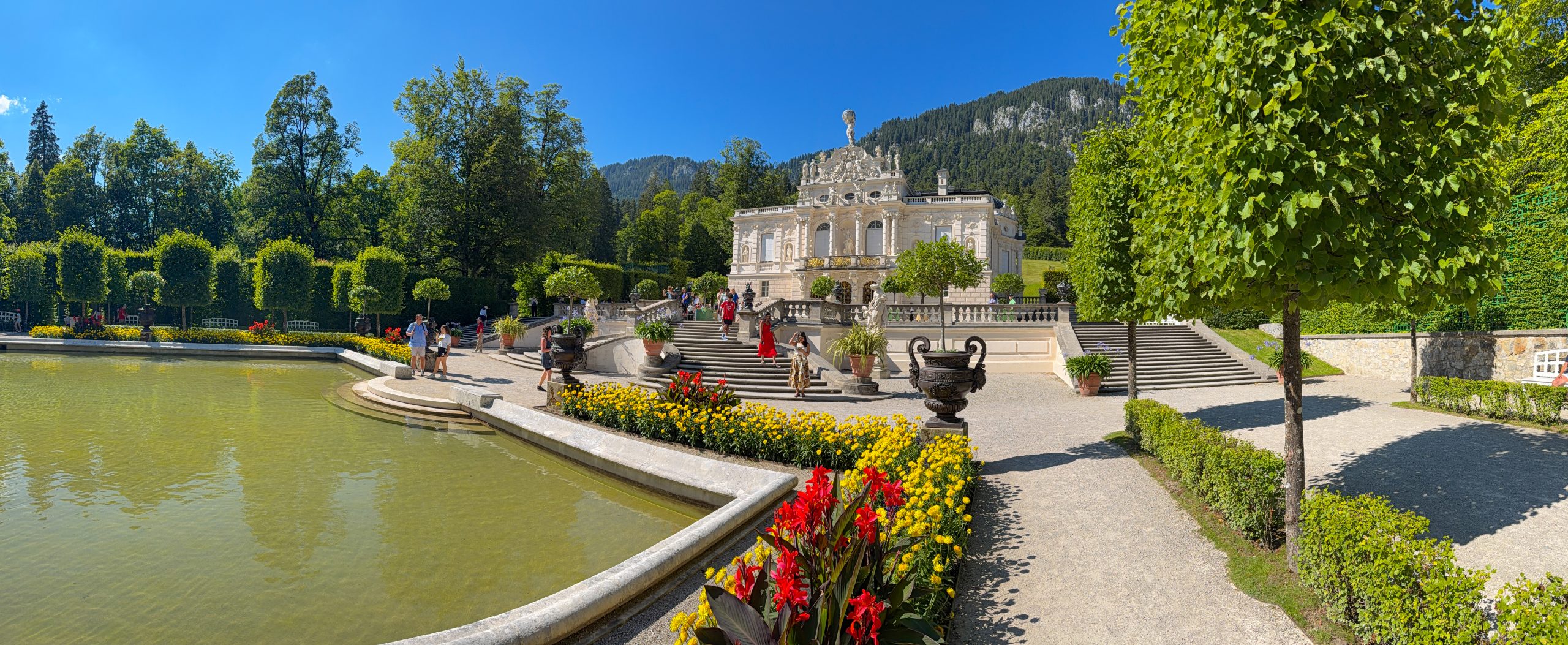 Elegant palace with gardens, fountain, and tourists under a blue sky.