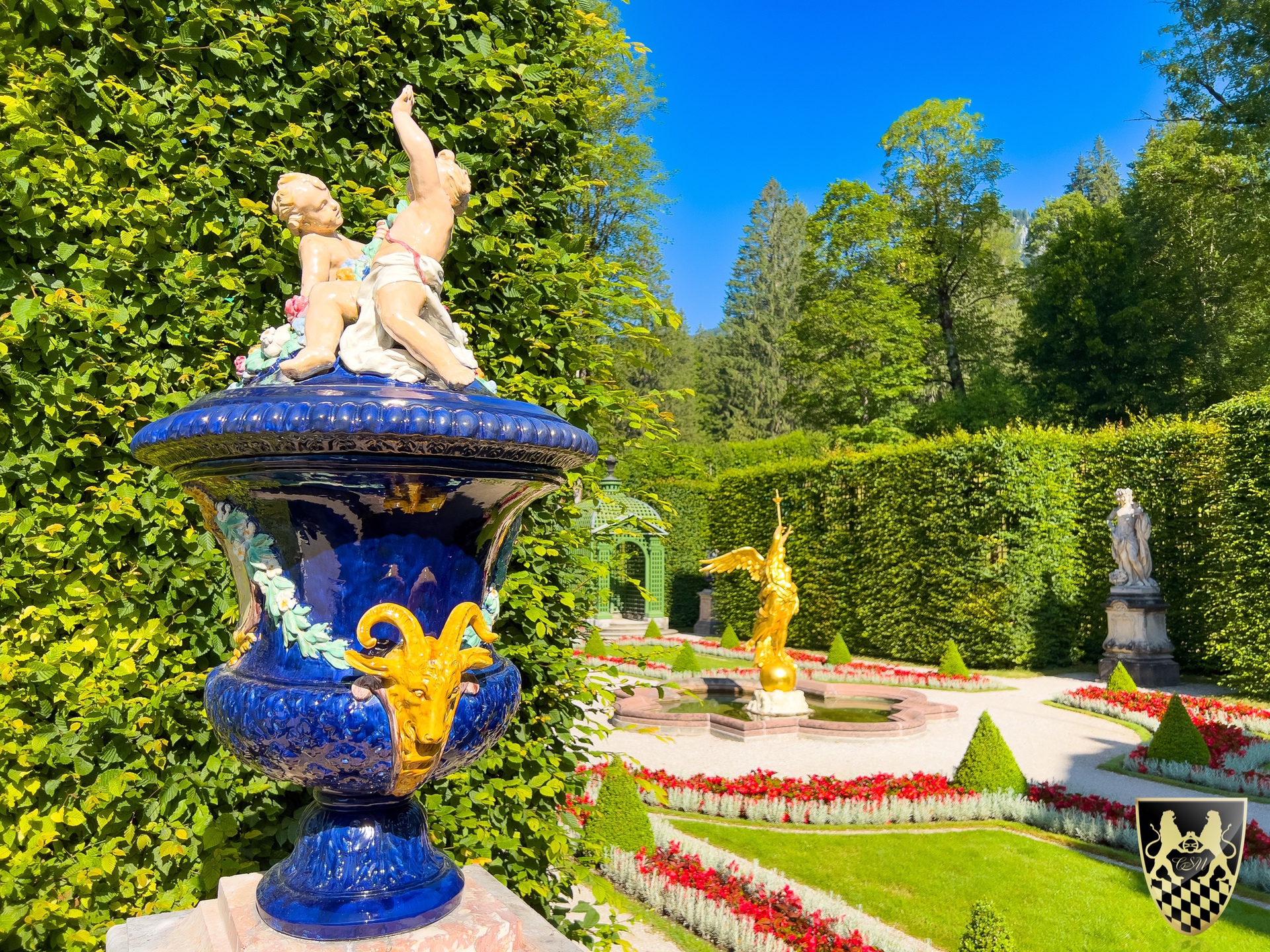 Ornate garden with blue vase, cherub statues, and manicured hedges.