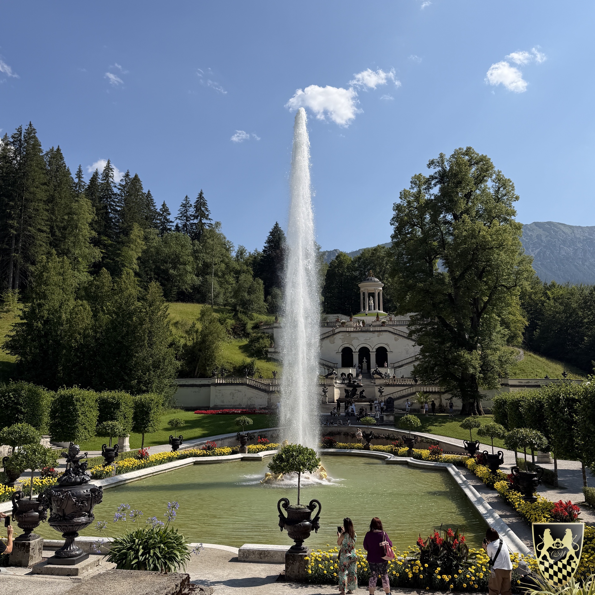 Fountain in garden with people, trees, and distant structure on a sunny day.