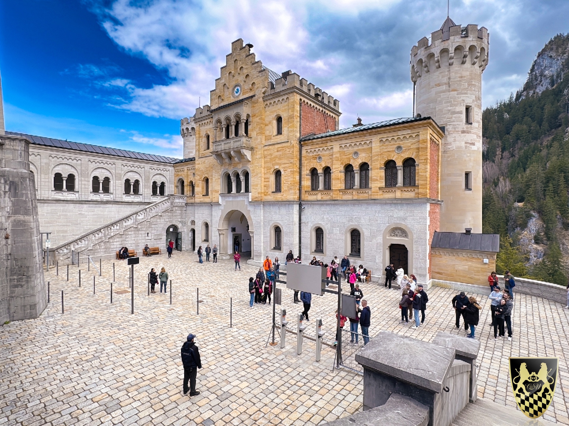 People in a courtyard of a historic European castle with towers and cloudy sky.